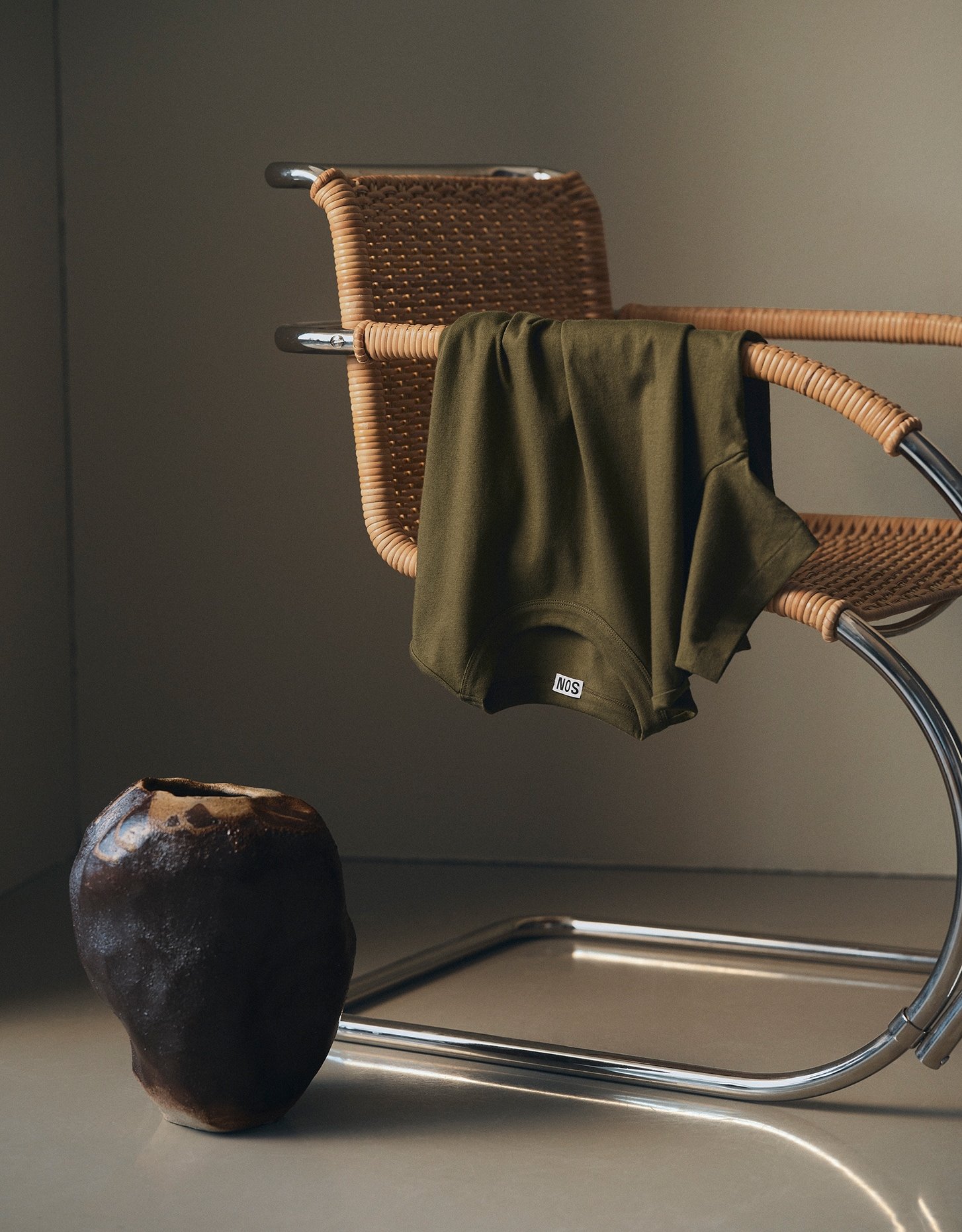 Olive green T-Shirt draped over a rattan chair beside a dark ceramic vase