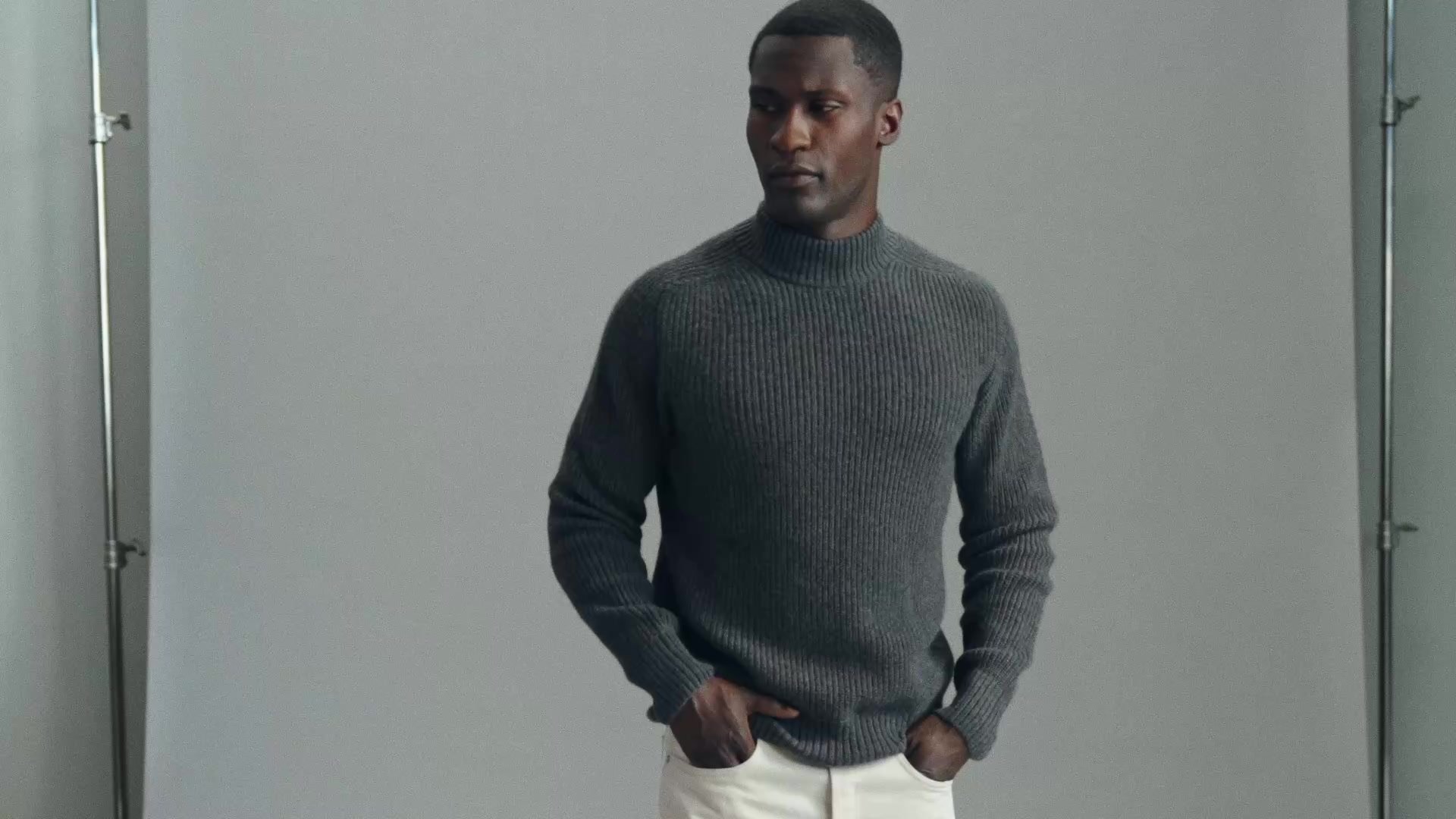 Model in a grey ribbed mock-neck sweater and off-white trousers, hands in pockets