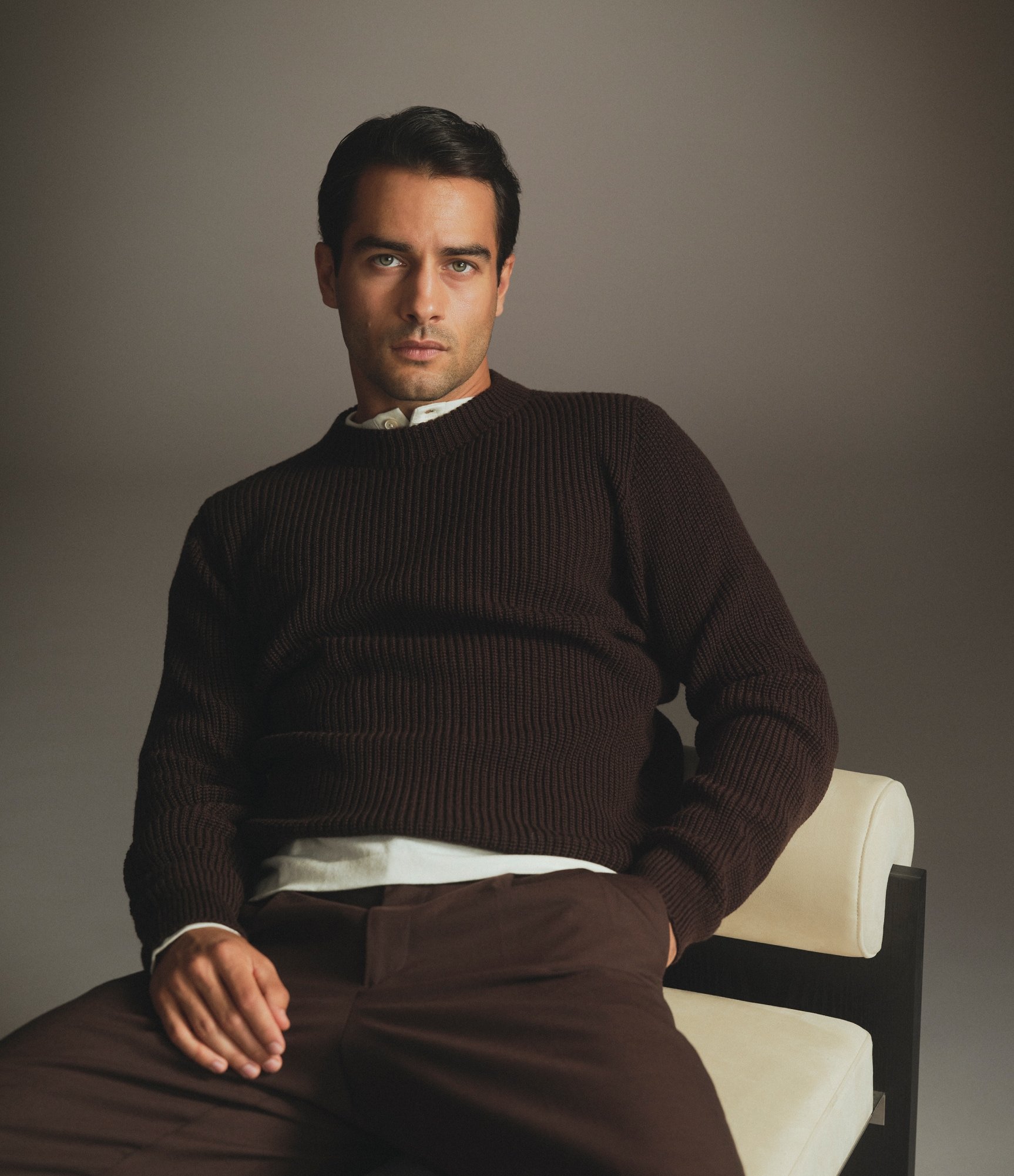 Model sat down wearing our heavy merino sweater in night brown