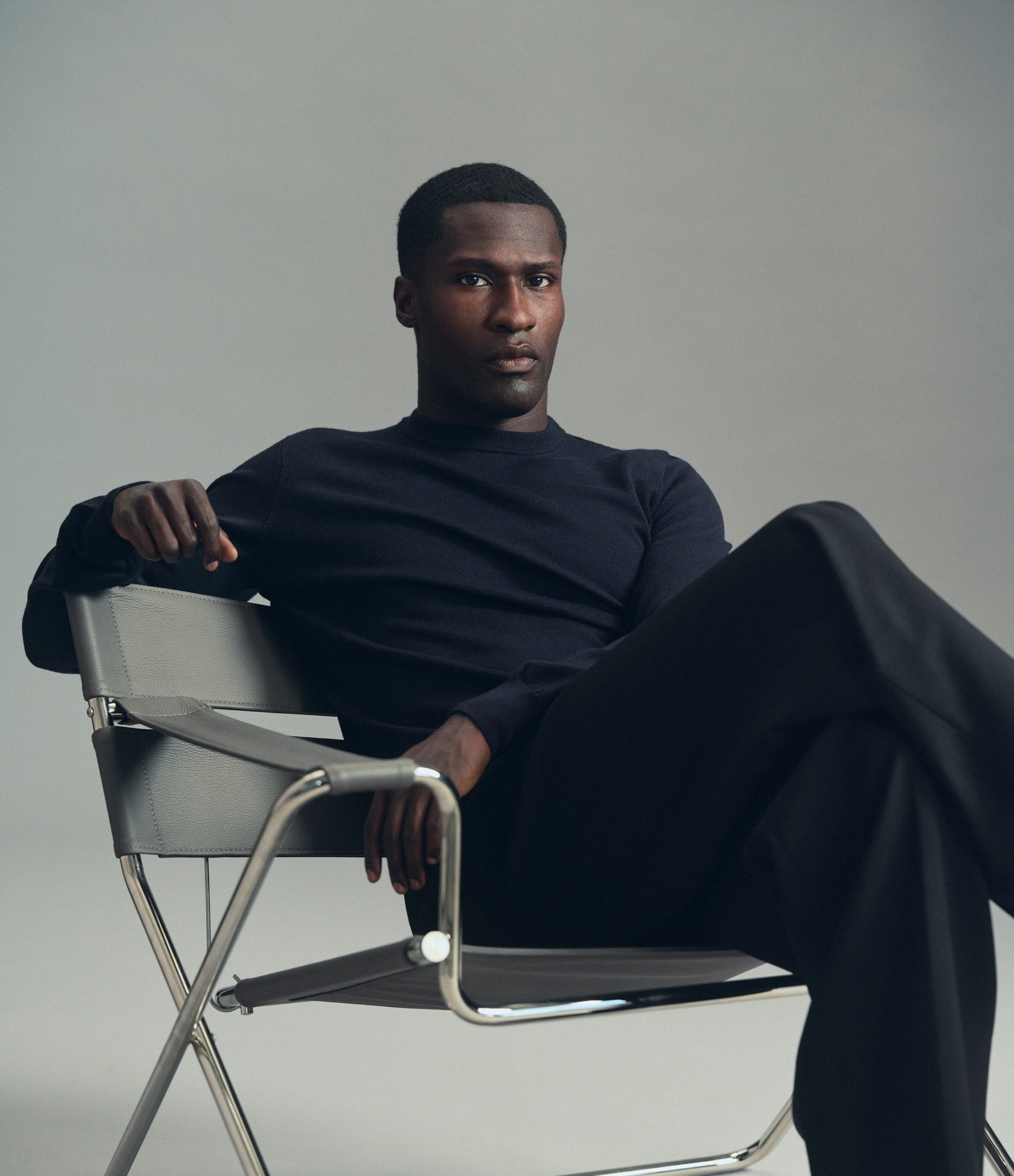Model sat on a leather chair wearing our merino pullover in navy